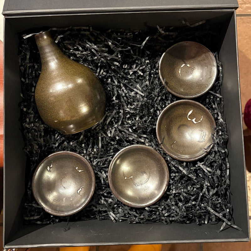 Handmade stoneware sake set by Kazan Studio featuring one sculptural tokkuri bottle and four guinomi cups with a dark metallic olive glaze, nestled in black shredded paper inside a matte black gift box